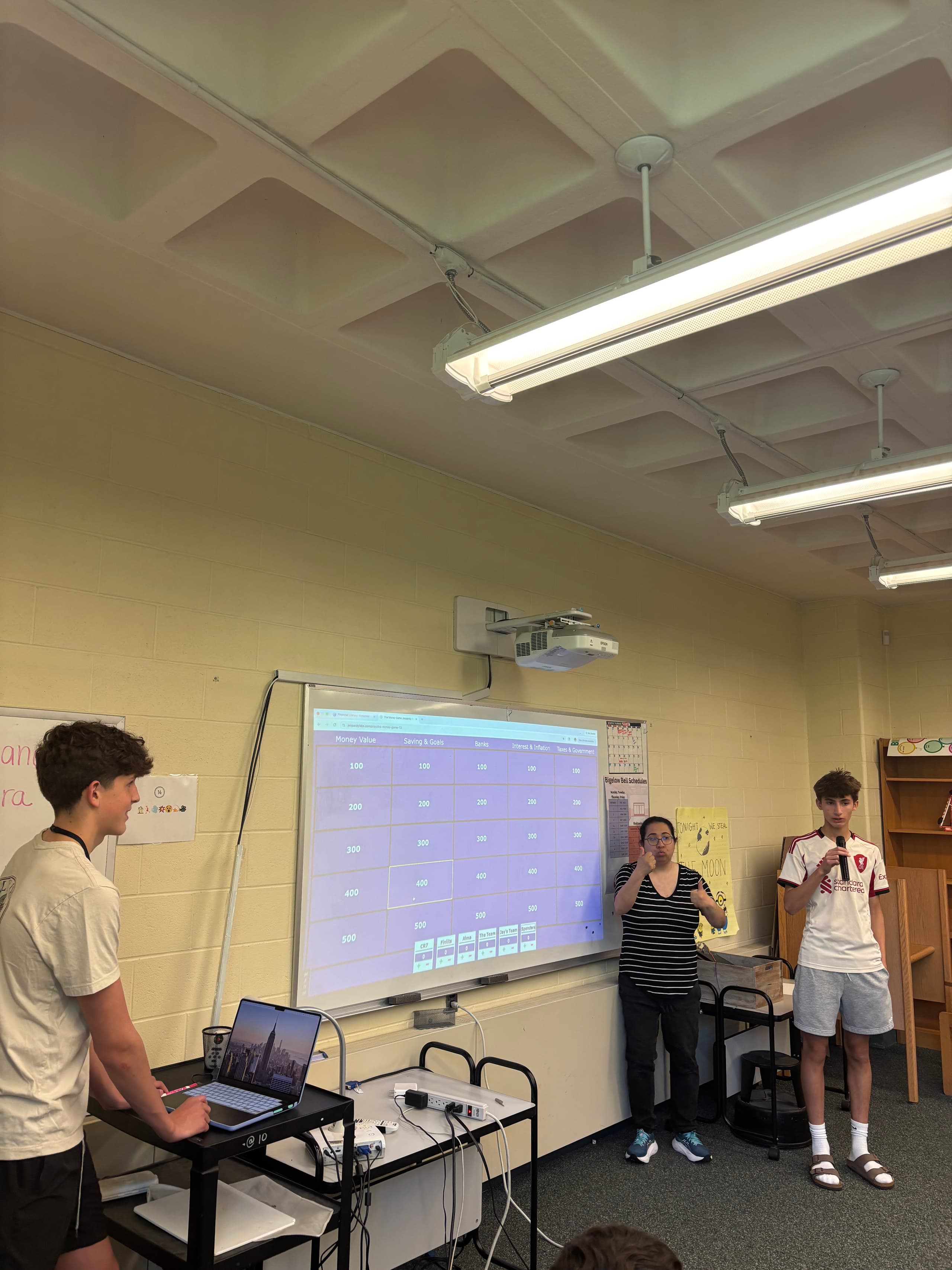 Compass presenters running a money-themed Jeopardy review game with eighth graders, the board projected on screen.